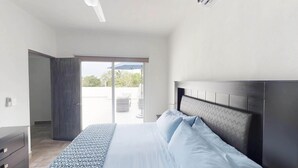 Ein Schlafzimmer mit einem Bett, einem dunklen Kopfteil, einem Nachttisch und Blick ins Freie durch eine Glastür.