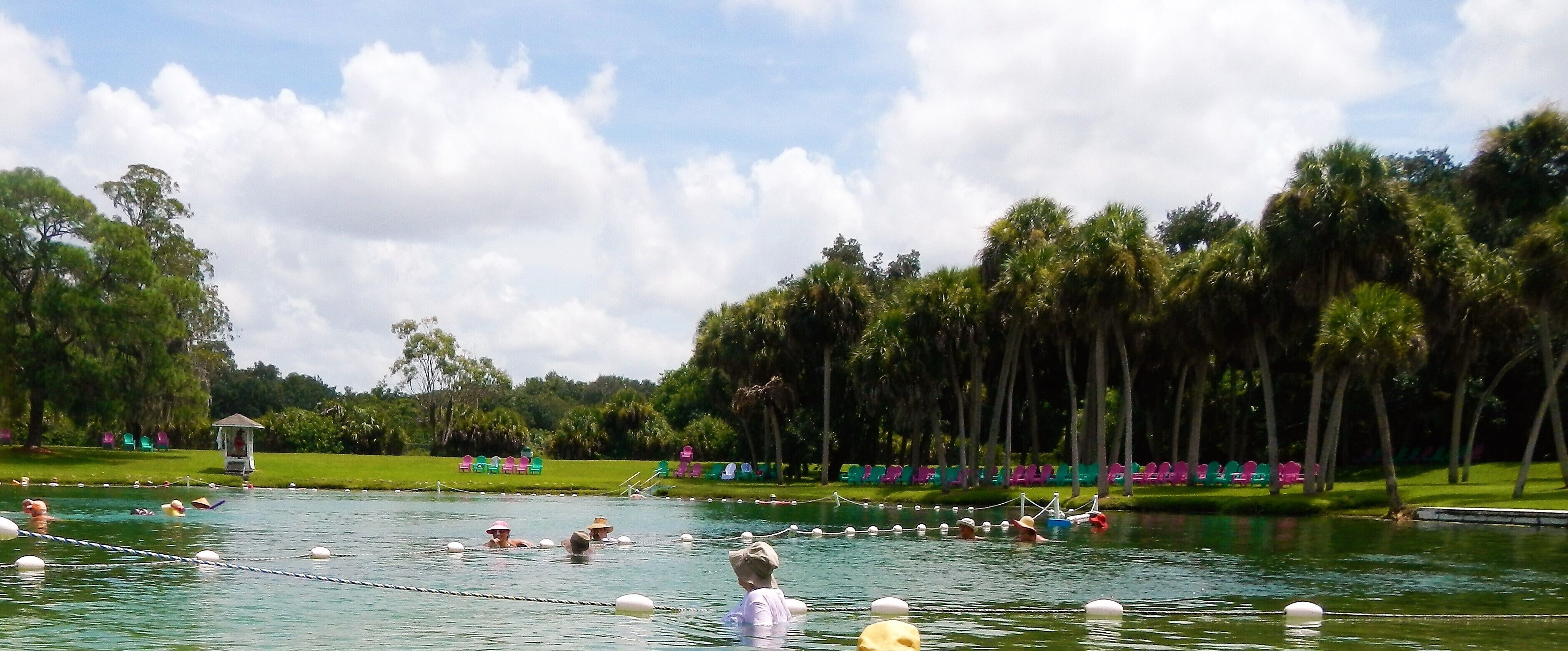 A swimming area with people in the water, surrounded by trees and a grassy area with colorful chairs.