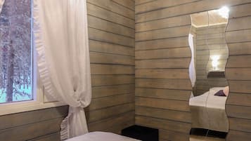 A bedroom with wooden panel walls, a bed with white bedding, a window with a curtain, and a mirror with a wavy frame.