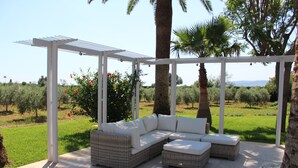 Un espacio de estar al aire libre cubierto con una pérgola blanca, mobiliario de mimbre y vistas a un paisaje verde.