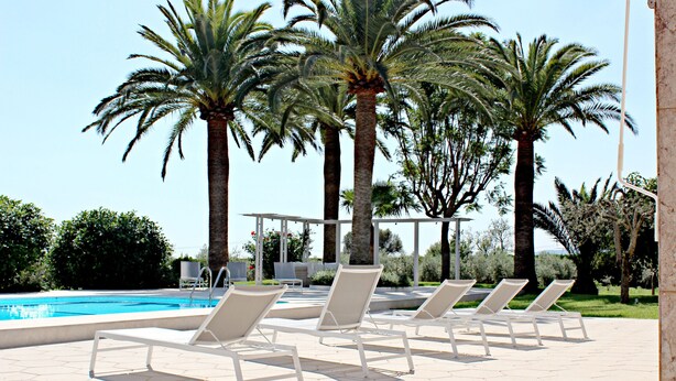Un área junto a la piscina con sillones de descanso y palmeras.