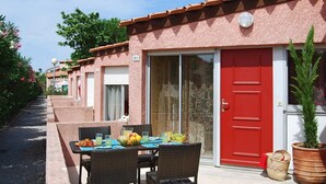 Una hilera de edificios rosas con puertas rojas, con los números 312 y 314, que cuenta con mesas al aire libre con sillas de mimbre y una disposición de frutas y bebidas.