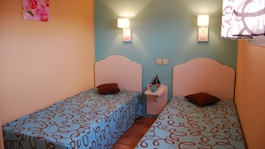 Ein Zimmer mit zwei Betten, jeweils mit einem gemusterten Überwurf und einem Kissen. An den Wänden sind Lampen angebracht und ein kleiner Tisch mit einem dekorativen Gegenstand steht daneben.