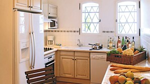 A kitchen with wooden cabinets, a refrigerator, a microwave, a sink, and a dining area with a bench and a table. - Maison Équipée + Climatisée | Accès piscine et spa (Fayence)