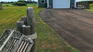 A green house with a white garage door, surrounded by a grassy field and a wooden fence. - Cavendish Driftwood Getaway - Beach, National Park, Nature Trails, Seafood, Golf (Cavendish)