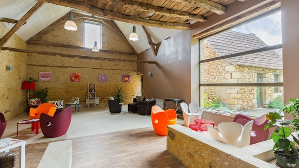 A spacious loft with exposed wooden beams, a stone wall, and a large window offering a view of a stone building outside.