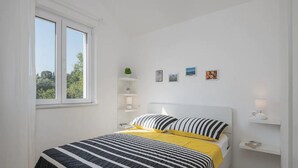 A bedroom with a bed, a window, a bedside table, and a shelf with decorative items.