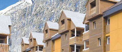 A row of wooden multi-story buildings with balconies, set against a snowy mountain backdrop.