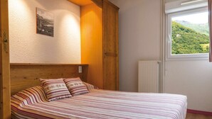 A bedroom with a wooden headboard, a bed with striped bedding, a window with a view of greenery, and a radiator. - Appart Équipé Près du Centre-Ville (Cauterets)