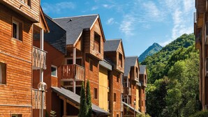 A row of wooden multi-story buildings with balconies, set against a backdrop of a mountain and lush greenery.