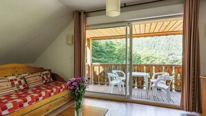 A cozy room with a wooden sofa, a small table, and a balcony with white plastic chairs and a wooden table.