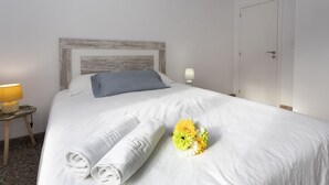 Ein ordentlich bezogenes Bett mit weißen Leinen, einem grauen Kopfkissen und einer Blumenkomposition.