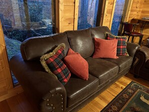Ein braunes Ledersofa mit Karokissen und einem Fellüberwurf, platziert in einer Holzhütte mit großen Fenstern.