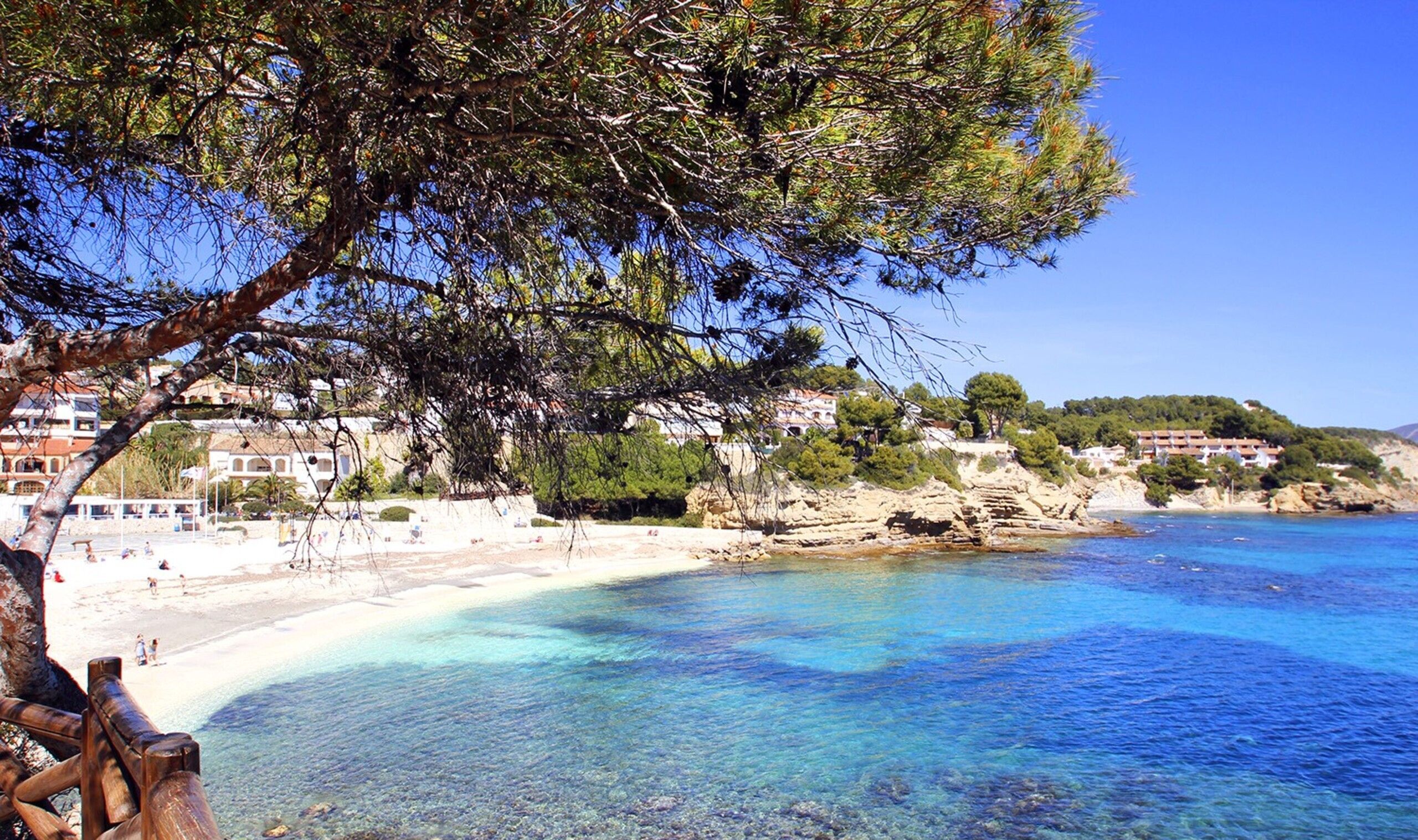 A coastal scene with clear turquoise waters, a sandy beach, and buildings in the background.