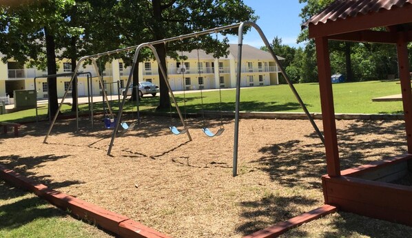 Un terrain de jeux avec des balançoires et un bac à sable, entouré d’un bâtiment et d’une zone herbeuse.