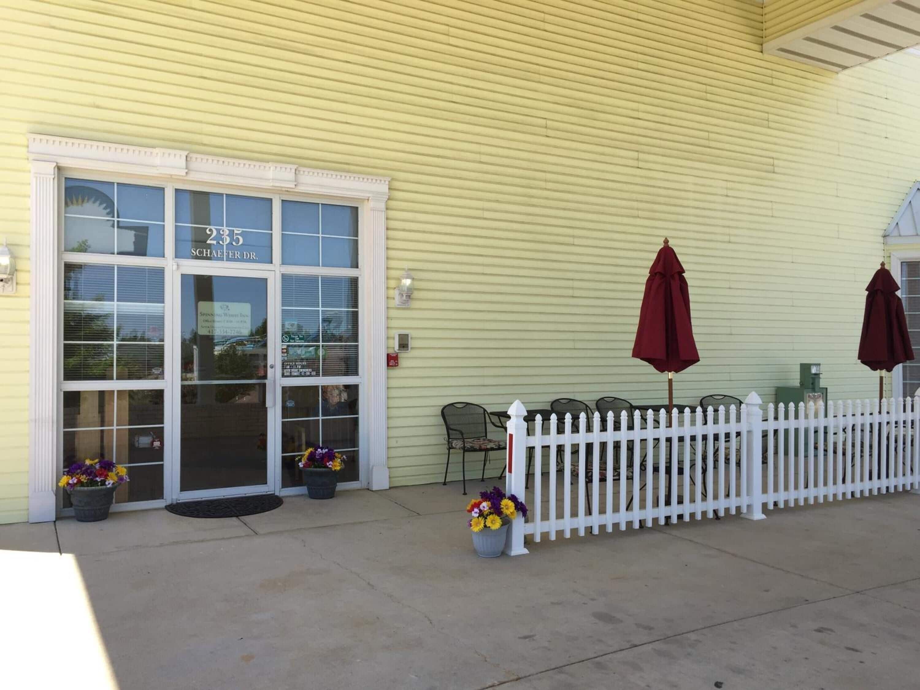 A yellow building with a white picket fence, outdoor seating with umbrellas, and flower pots.
