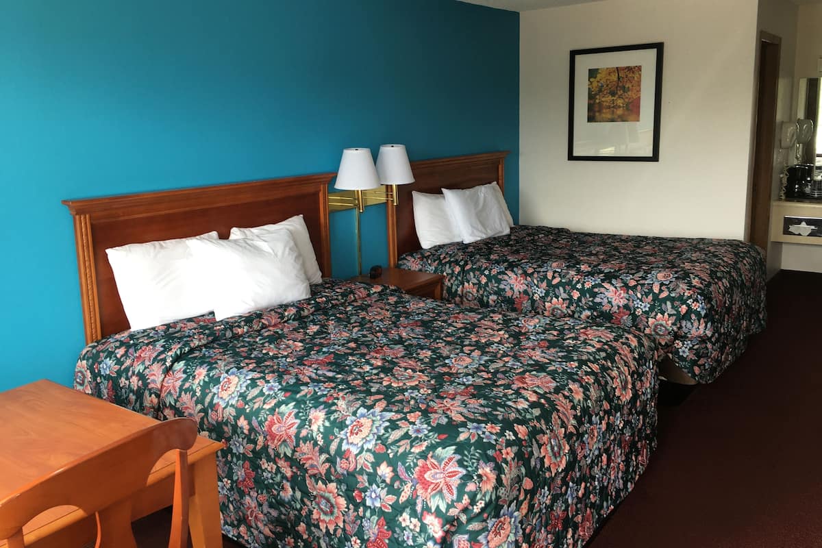 Une chambre d’hôtel avec deux lits simples, un bureau en bois, une chaise, une table de chevet avec des lampes et un tableau encadré au mur.