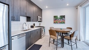 A modern kitchen with dark wood cabinets, stainless steel appliances, and a white countertop. There is a dining area with a round table and four chairs. A painting of a garden is on the wall.