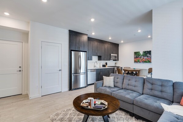 A modern living room with a grey sofa, a coffee table with books and a tray, a kitchen with dark cabinets and stainless steel appliances, and a painting on the wall.