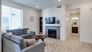 A modern living room with a grey sectional sofa, a round coffee table, a fireplace, a flat-screen TV, and a large window with blinds.