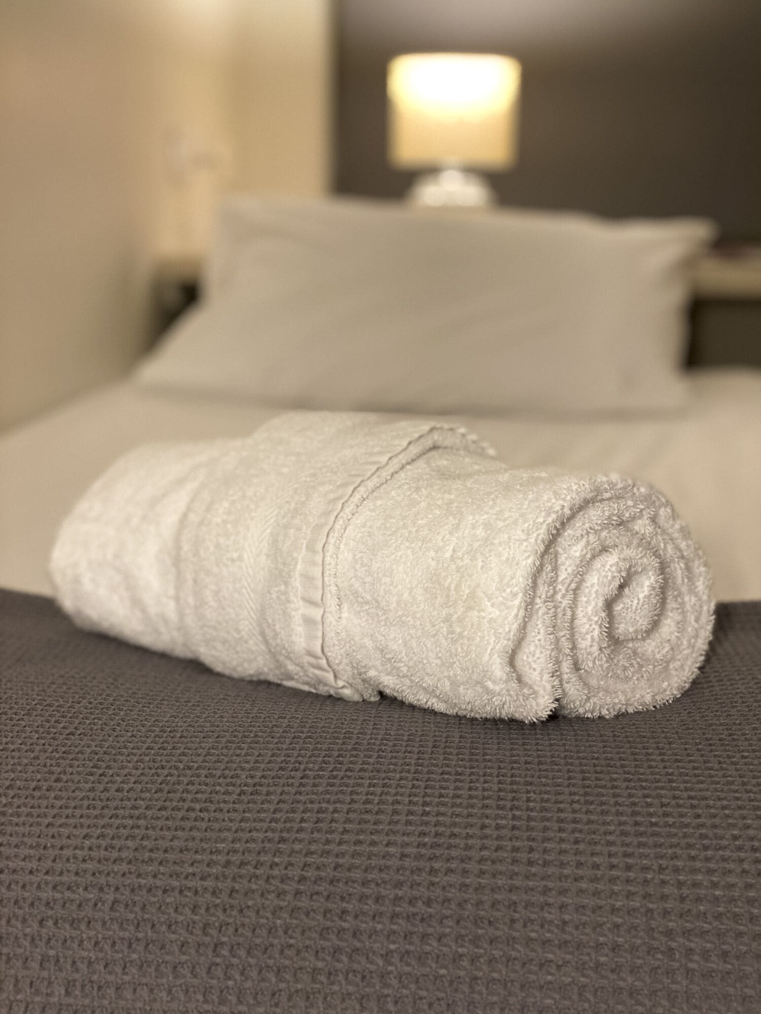 A neatly made bed with a white pillow and a rolled white towel on a textured gray bedspread.