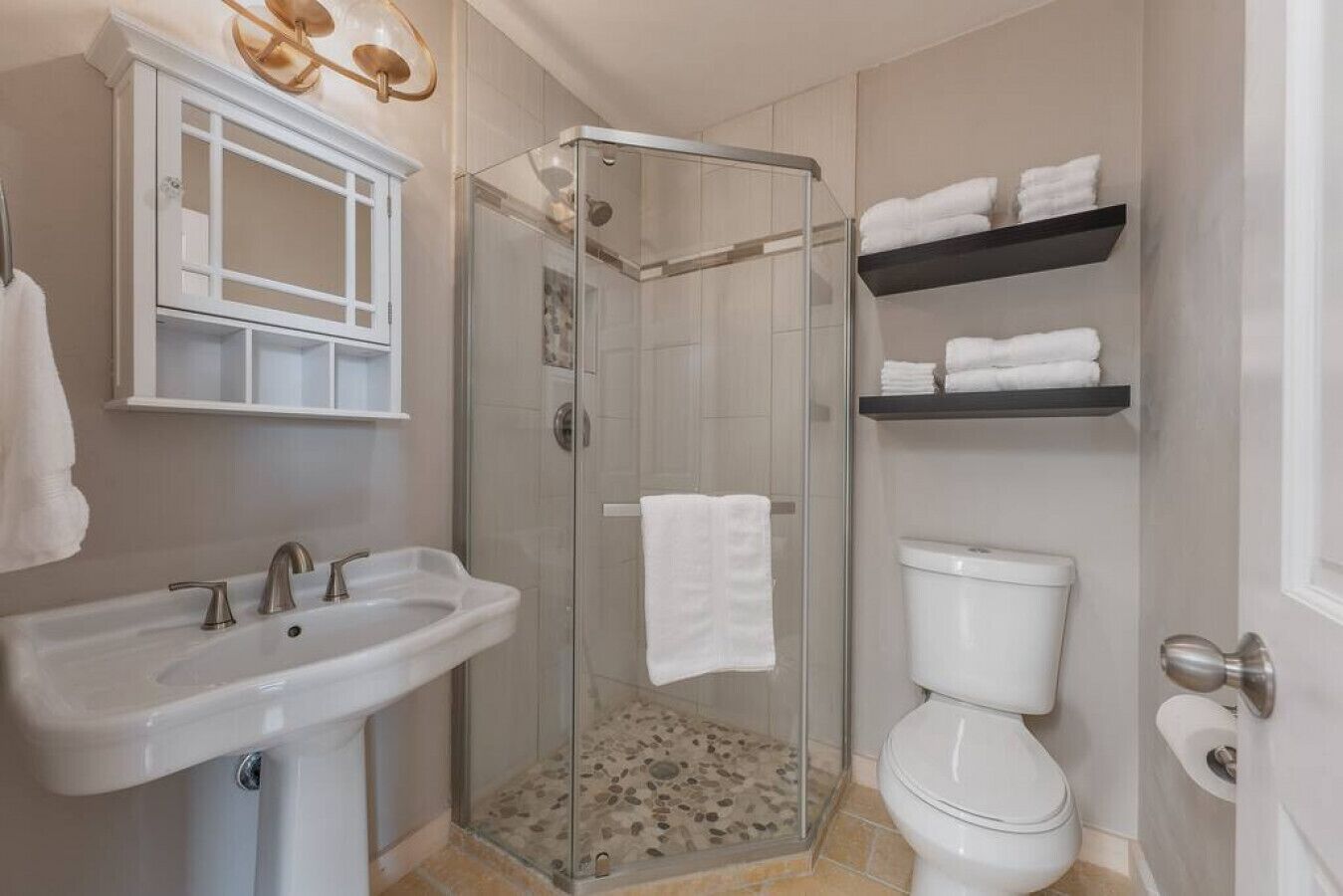 A modern bathroom with a glass-enclosed shower, a white sink, a toilet, and two shelves with folded towels.