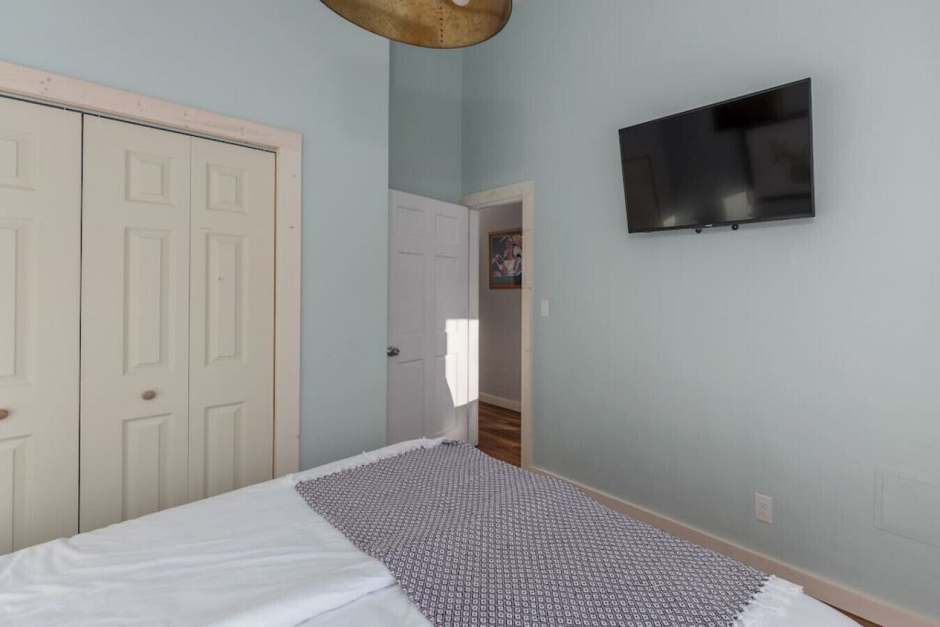 Un dormitorio con una cama, un televisor montado en la pared y una puerta que da a otra habitación.