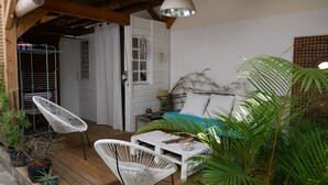 A cozy bedroom with a bed, two white wicker chairs, a small table, and a nightstand. There is a window with a curtain, a wooden ceiling, and a wall-mounted fan. - From the beach to golf (Saint-Gilles-les Bains)