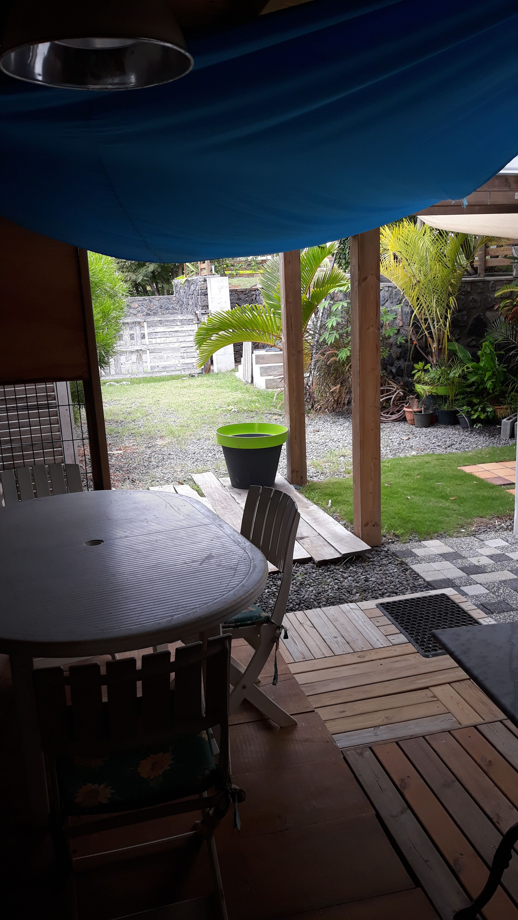 Ein überdachter Patio-Bereich mit einem runden Tisch und Stühlen, einem blauen Sonnensegel und Blick auf einen Garten mit einer Steinmauer und Pflanzgefäßen.