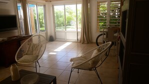 A room with two white wicker chairs, a TV, a table with a vase and a basket, and a sliding glass door leading to a garden. - From the beach to golf (Saint-Gilles-les Bains)