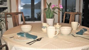 A dining table set with white ceramic dishes, a vase with pink flowers, and a view of the ocean through large windows. - Eagles Watch Ocean Front B and B, Auke Bay, Juneau
 CBJ 1000195 (Juneau)