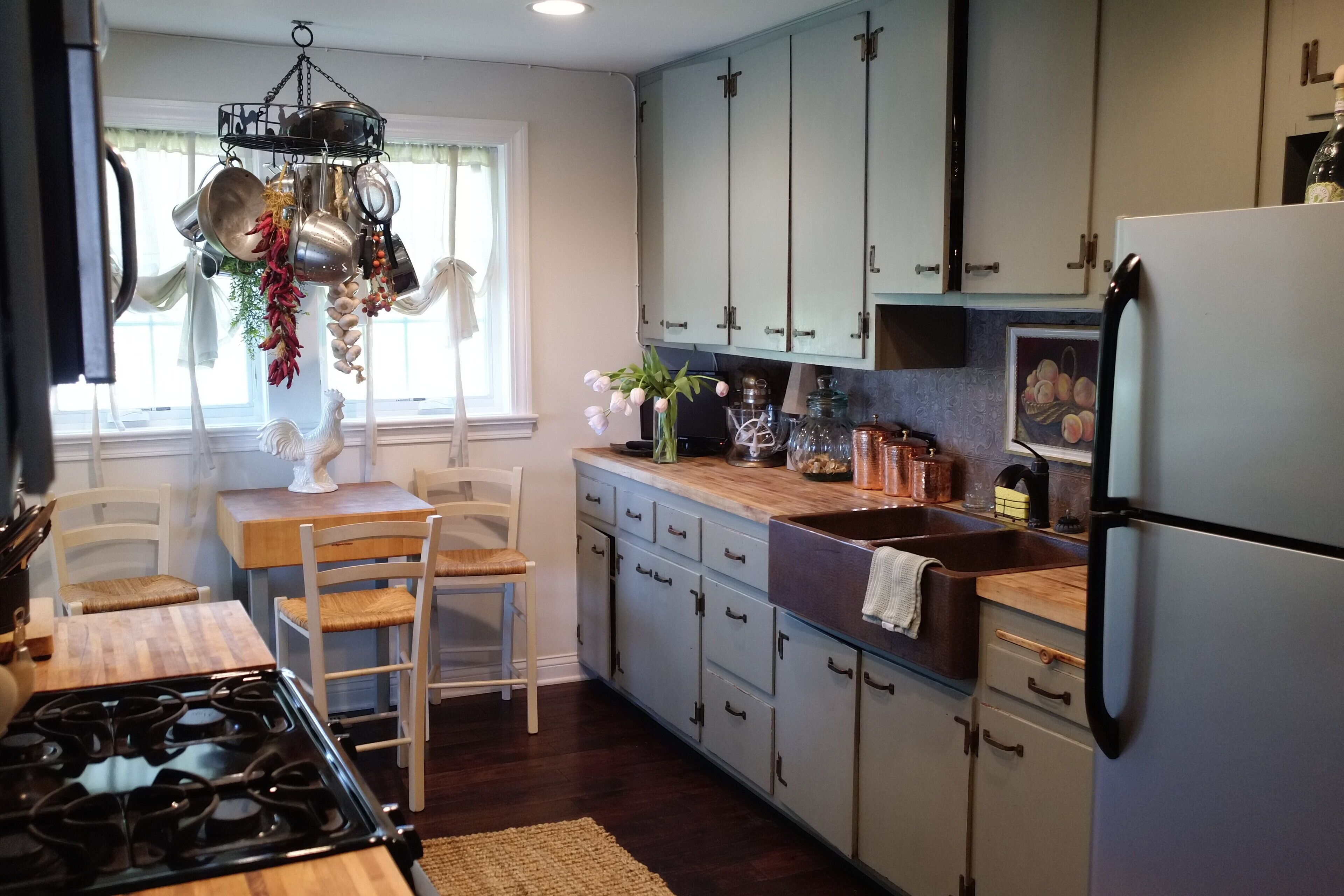 A kitchen with a stove, sink, and cabinets. There is a window with curtains and a hanging pot rack.