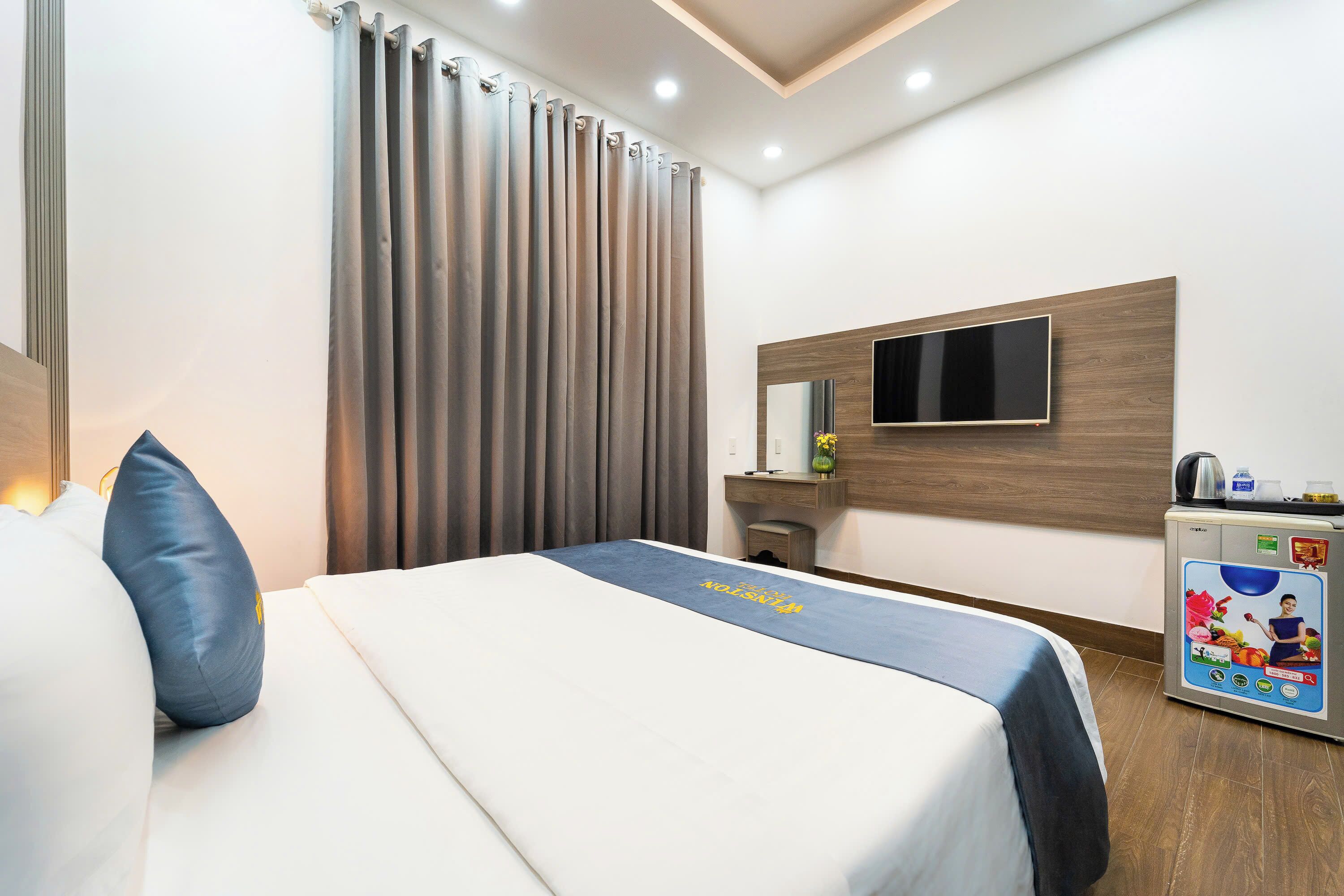 A hotel room with a bed, a TV mounted on the wall, a minibar, and a curtain.