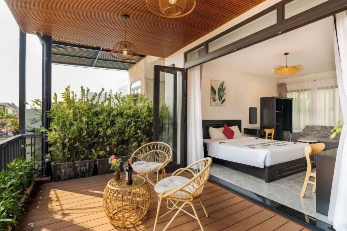 A modern bedroom with a large bed, a small seating area, and a balcony with plants.
