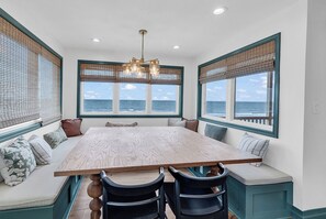 A dining room with a large table, chairs, and a view of the ocean through large windows.