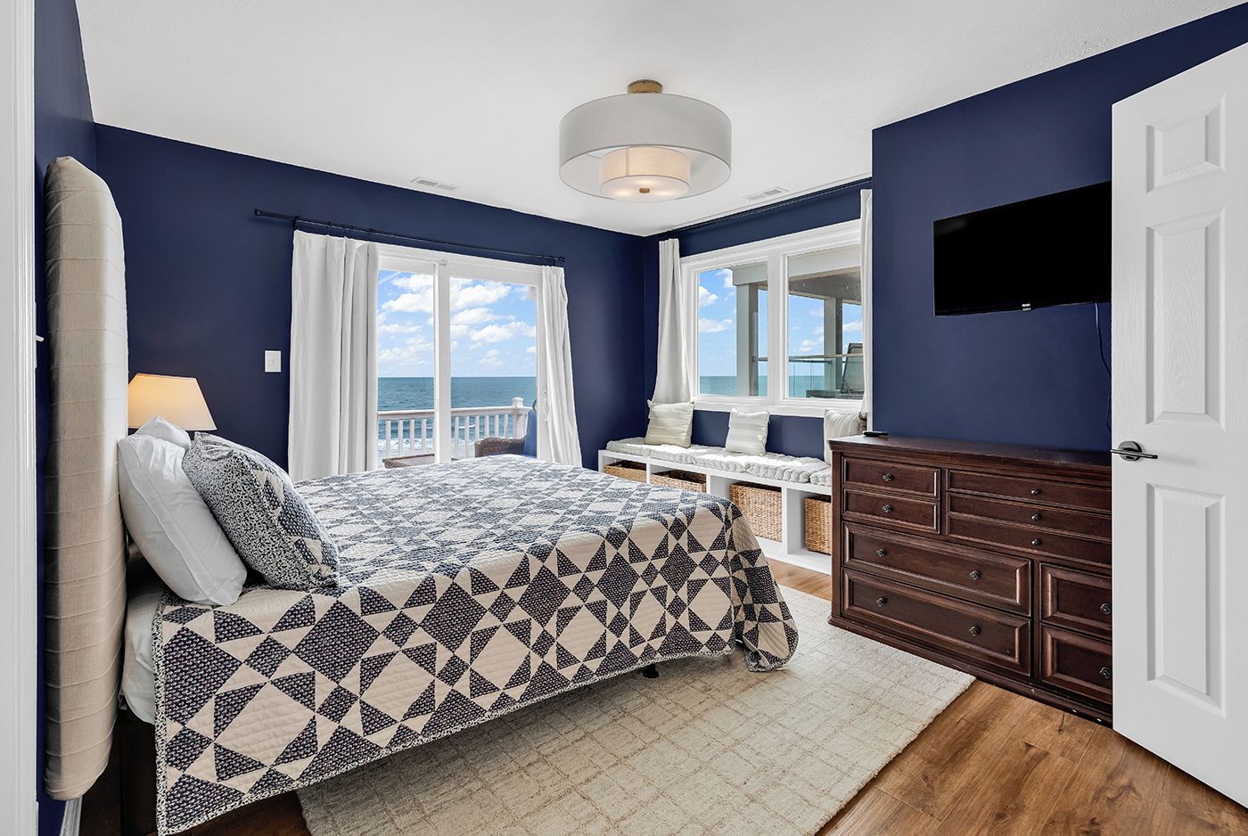 Een slaapkamer met een bed, een kaptafel, een raam met uitzicht op zee en een plafondlamp.