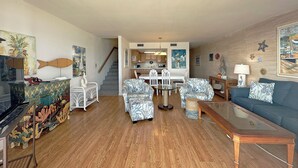 A living room with a wooden floor, a blue sofa, patterned armchairs, a glass coffee table, a dining table with chairs, and a kitchen in the background. - Shores of Surfside I 103 - Oceanfront in Surfside Beach (Surfside Beach)