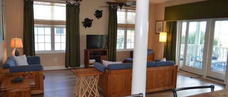 A spacious living room with a wooden floor, a ceiling fan, a television, and a balcony with a view.