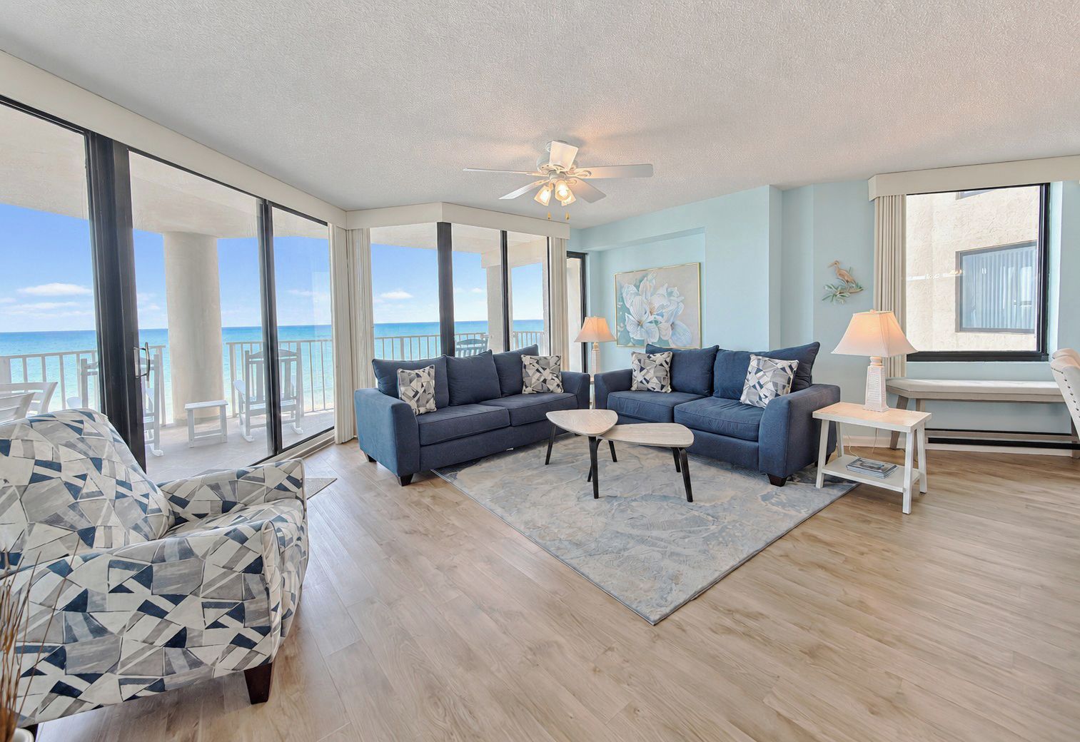 A spacious living room with a large sofa, a coffee table, and a chair. There is a balcony with a view of the ocean.