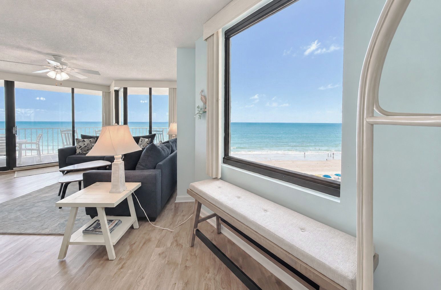 Ein Zimmer mit großem Fenster und Meerblick, Deckenventilator, Sofa, Lampe und ein kleiner Tisch.