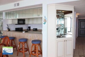 Cocina con armarios blancos, microondas y cafetera. Hay tres taburetes de madera con cojines azules.