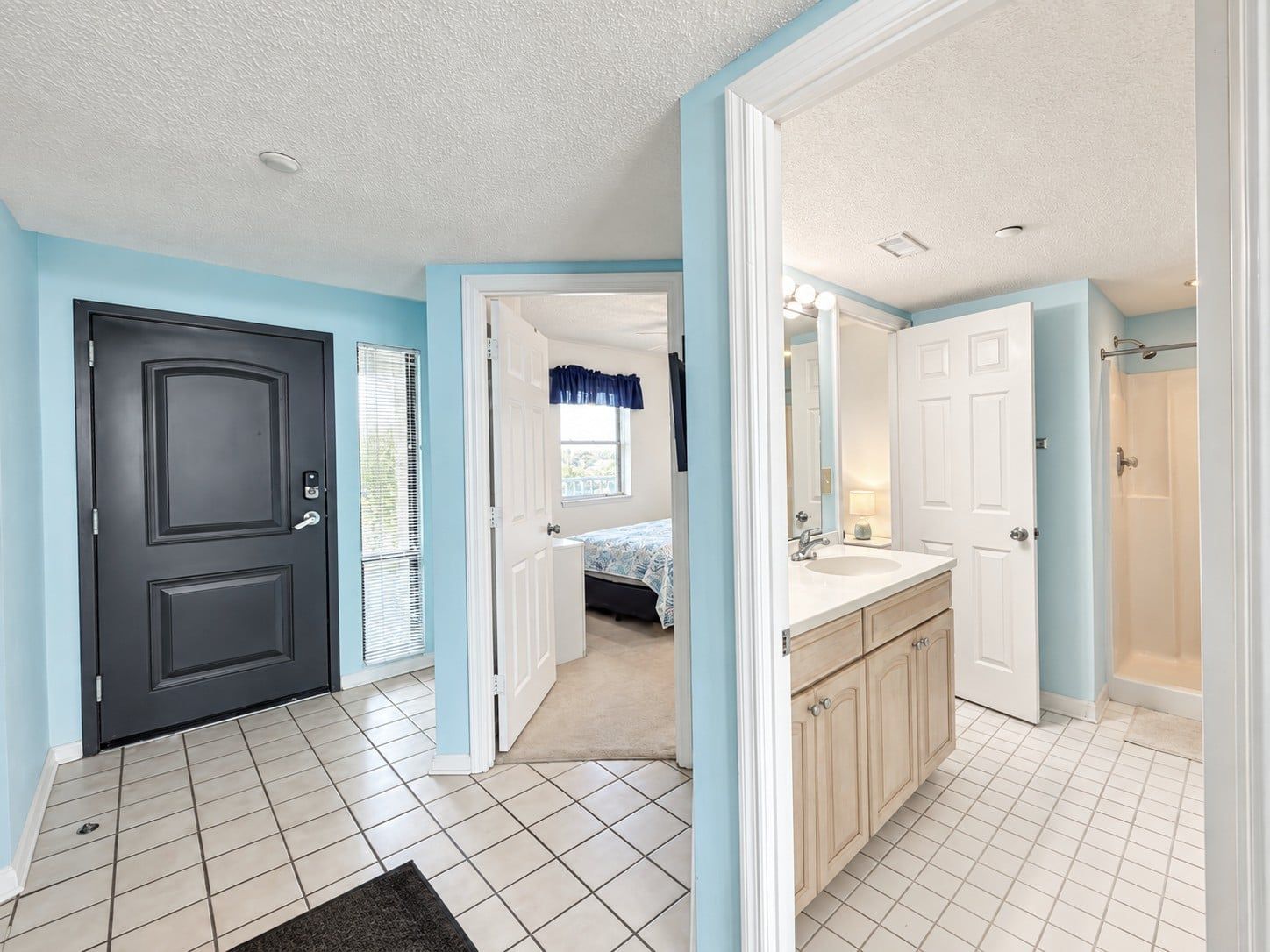 A hotel room with a door, a bed, a bathroom with a sink and mirror, and a view of the outdoors.