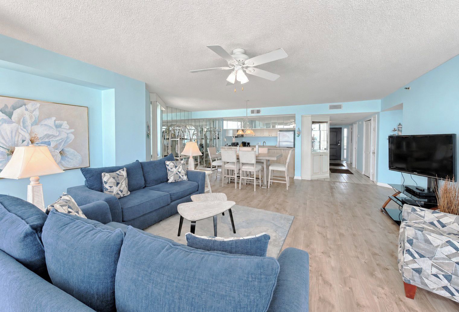 A living room with a blue sofa, a coffee table, and a TV. There is a ceiling fan and a dining area in the background.
