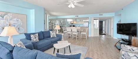 A living room with a blue sofa, a coffee table, and a TV. There is a ceiling fan and a dining area in the background.