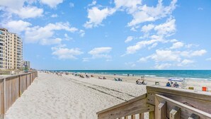 A beach with a wooden boardwalk leading to the shore, a sandy beach, and a building in the background. - Mariner's Watch 204 - Oceanfront in Garden City Beach (Garden City)