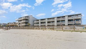 A beachfront hotel with multiple balconies, a sandy beach in the foreground, and a clear sky. - Mariner's Watch 204 - Oceanfront in Garden City Beach (Garden City)