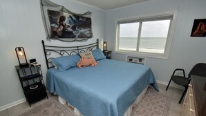 A bedroom with a bed, a nightstand, a lamp, a window with a view of the sea, and a wall-mounted artwork. - Sea Oaks 304 - Oceanfront in Garden City Beach (Garden City)