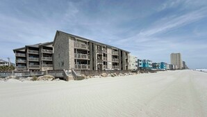 Ein Hotel direkt am Strand mit mehreren Balkonen, ein Sandstrand im Vordergrund und ein klarer Himmel.