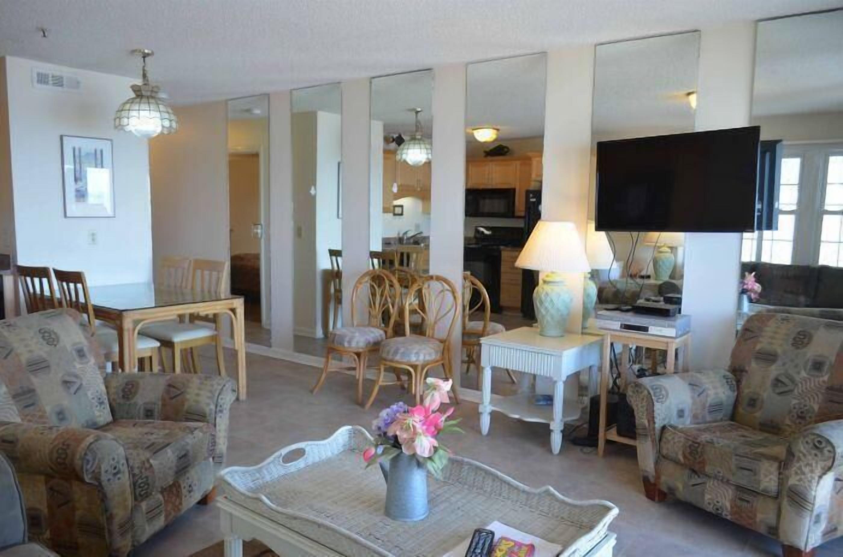 A living room with patterned armchairs, a dining table with chairs, a TV, and a kitchen in the background.
