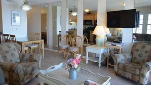 Een woonkamer met gestreepten fauteuils, een eettafel met stoelen, een televisie en een keuken op de achtergrond.